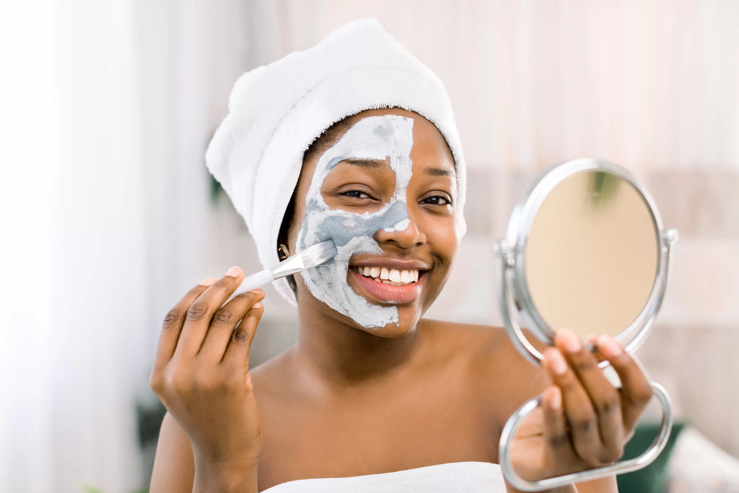 Young Black lady applying facial mask while looking in a hand mirror and smiling