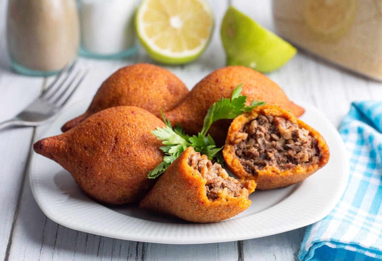 A plateful of kibbeh with lemons and a jar of drink in the background