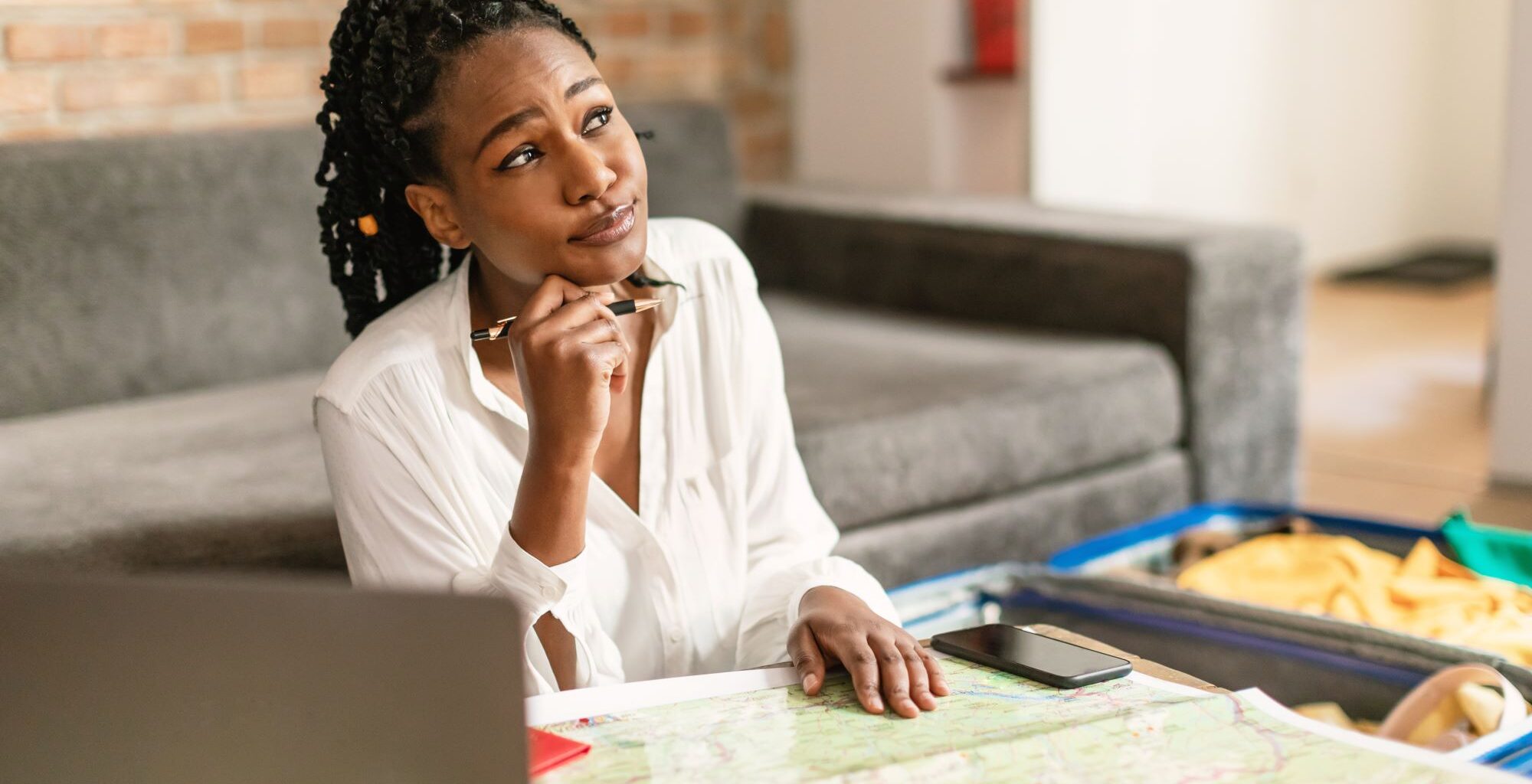 Dreamy African American woman planning future journey with map and laptop, thinking about best travel insurance essentials