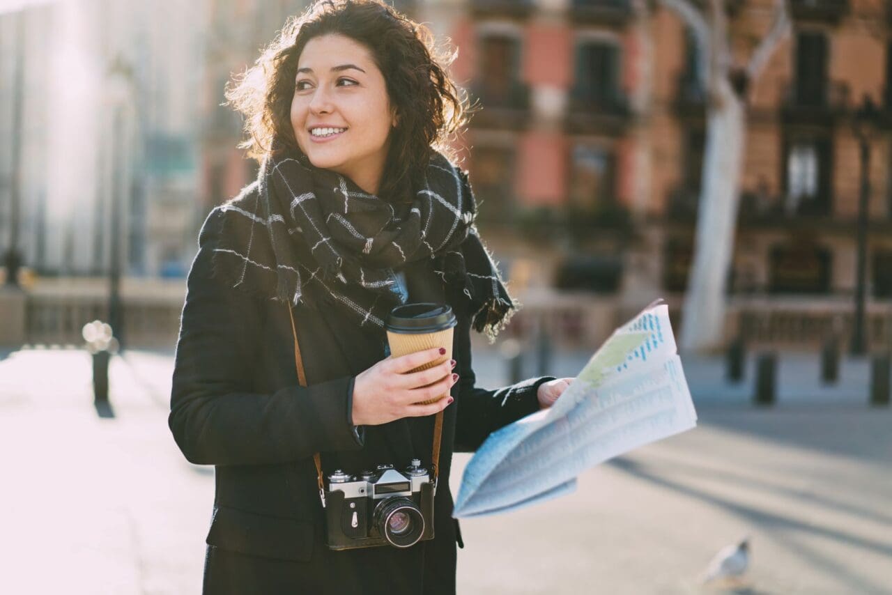 Cheerful tourist in stylish outfit holding coffee to go and location map and smiling